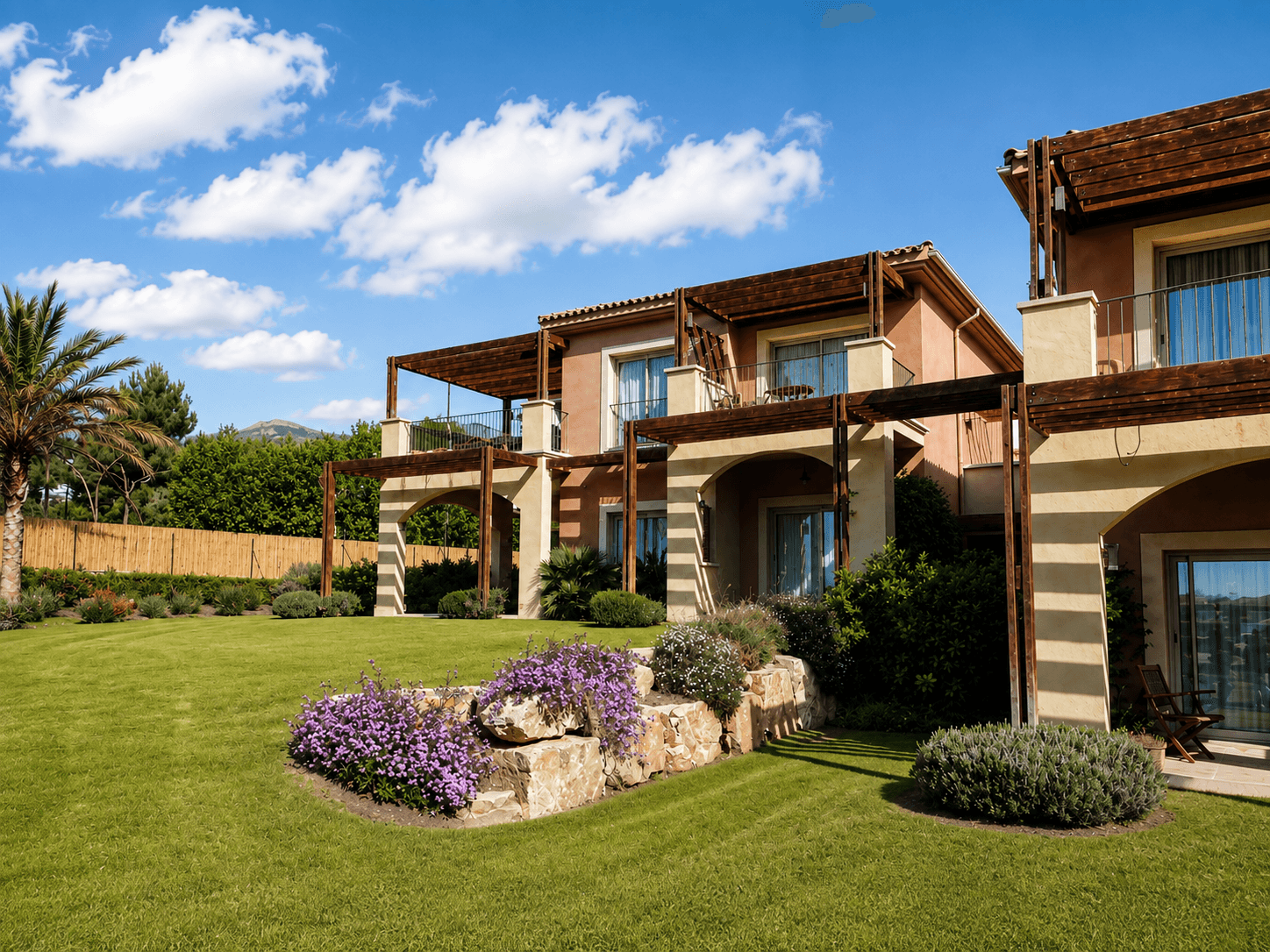 Viviendas de dos plantas con porches de madera y esquinas de piedra, sobre césped con macizos de lavanda y una palmera.
