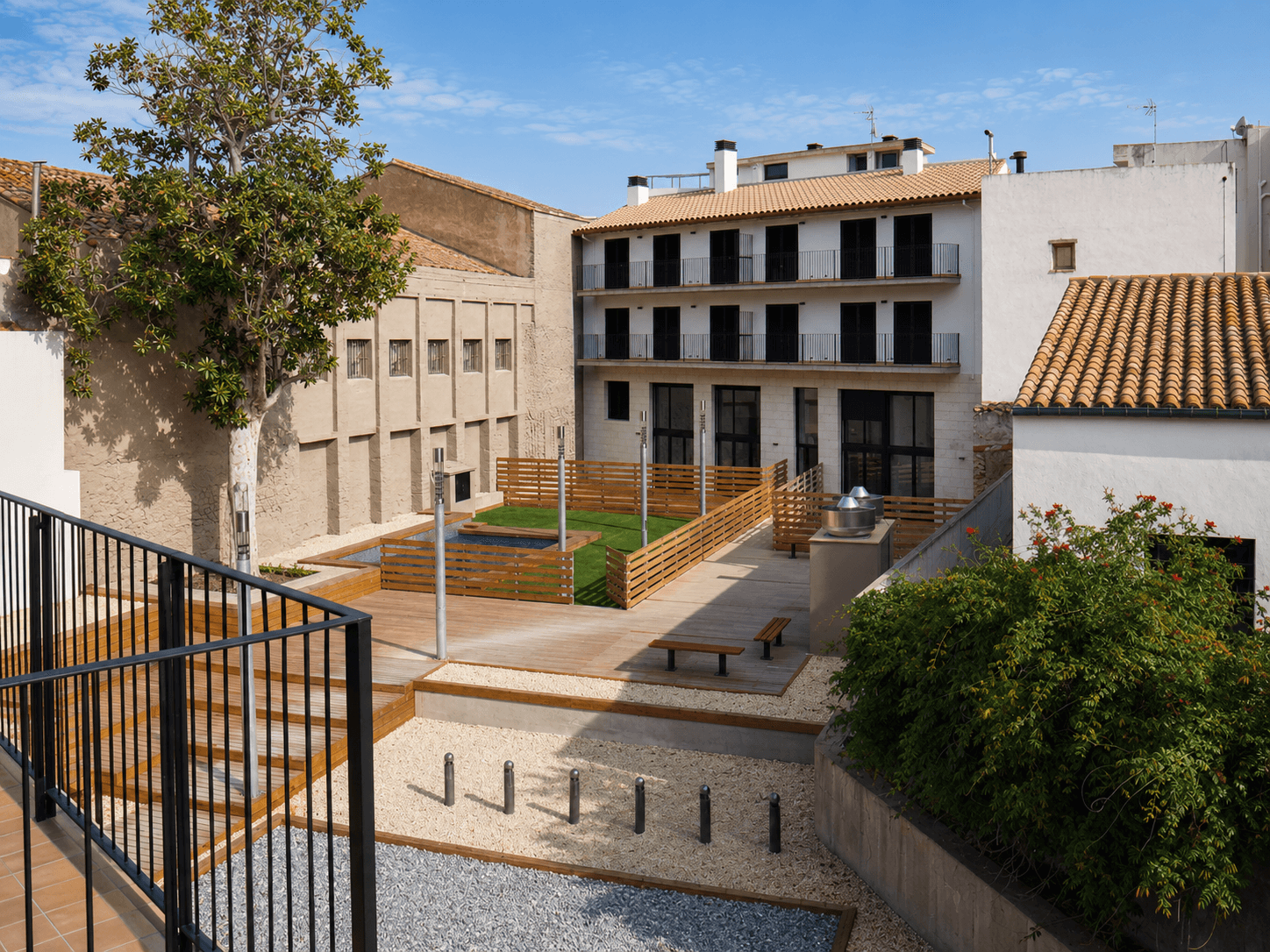 Patio interior de manzana con un edificio blanco de persianas oscuras, tarima de madera, gradas y bancos de obra.
