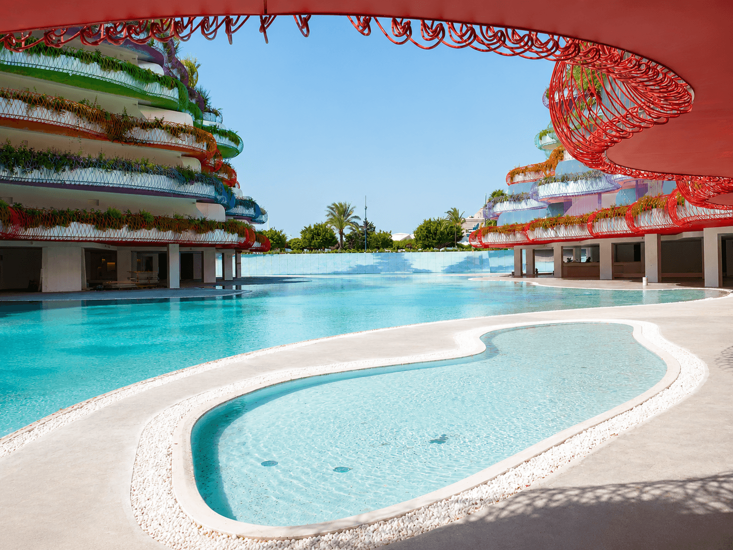 Piscina de trazado orgánico entre torres de balcones circulares de colores, con una celosía roja en primer término.