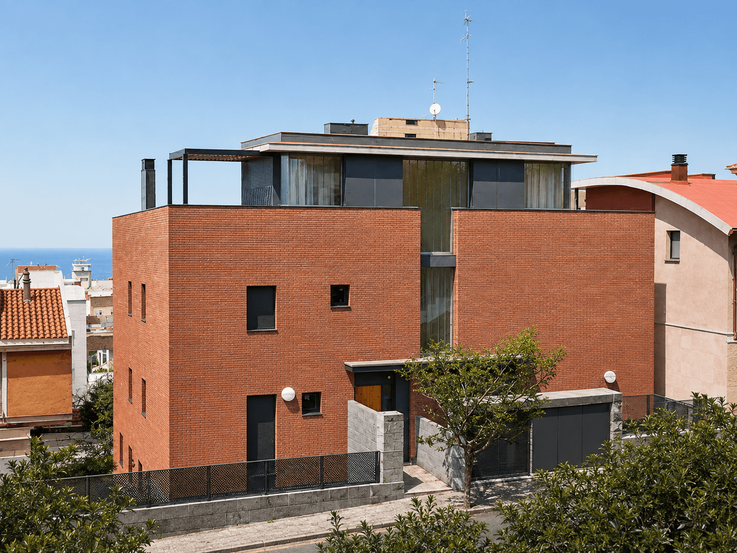 Edificio de ladrillo rojo de dos plantas con un ático acristalado retranqueado y el mar al fondo.