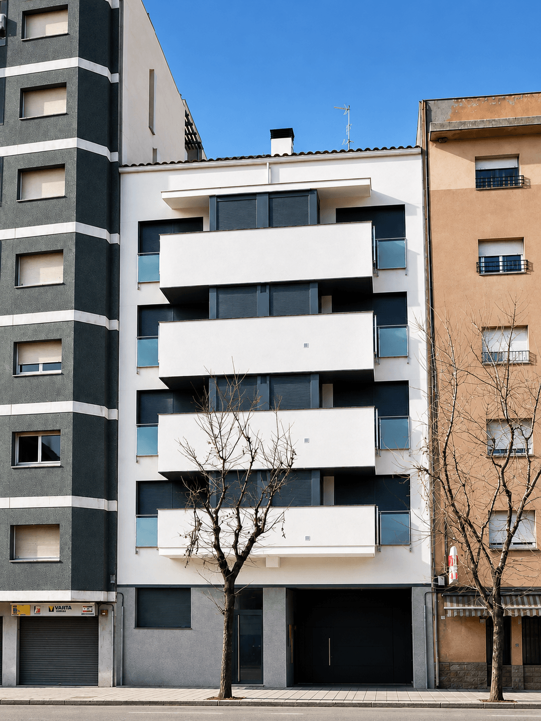 Edificio de cinco plantas entre medianeras, de fachada blanca con balcones corridos y carpintería oscura, con dos árboles sin hoja delante.