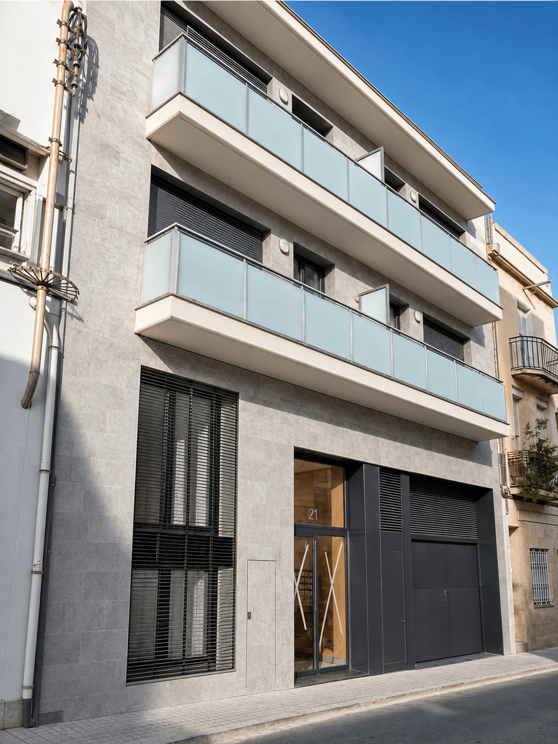 Edificio entre medianeras de fachada de piedra gris, con balcones de vidrio, portal de madera y puerta de garaje oscura.