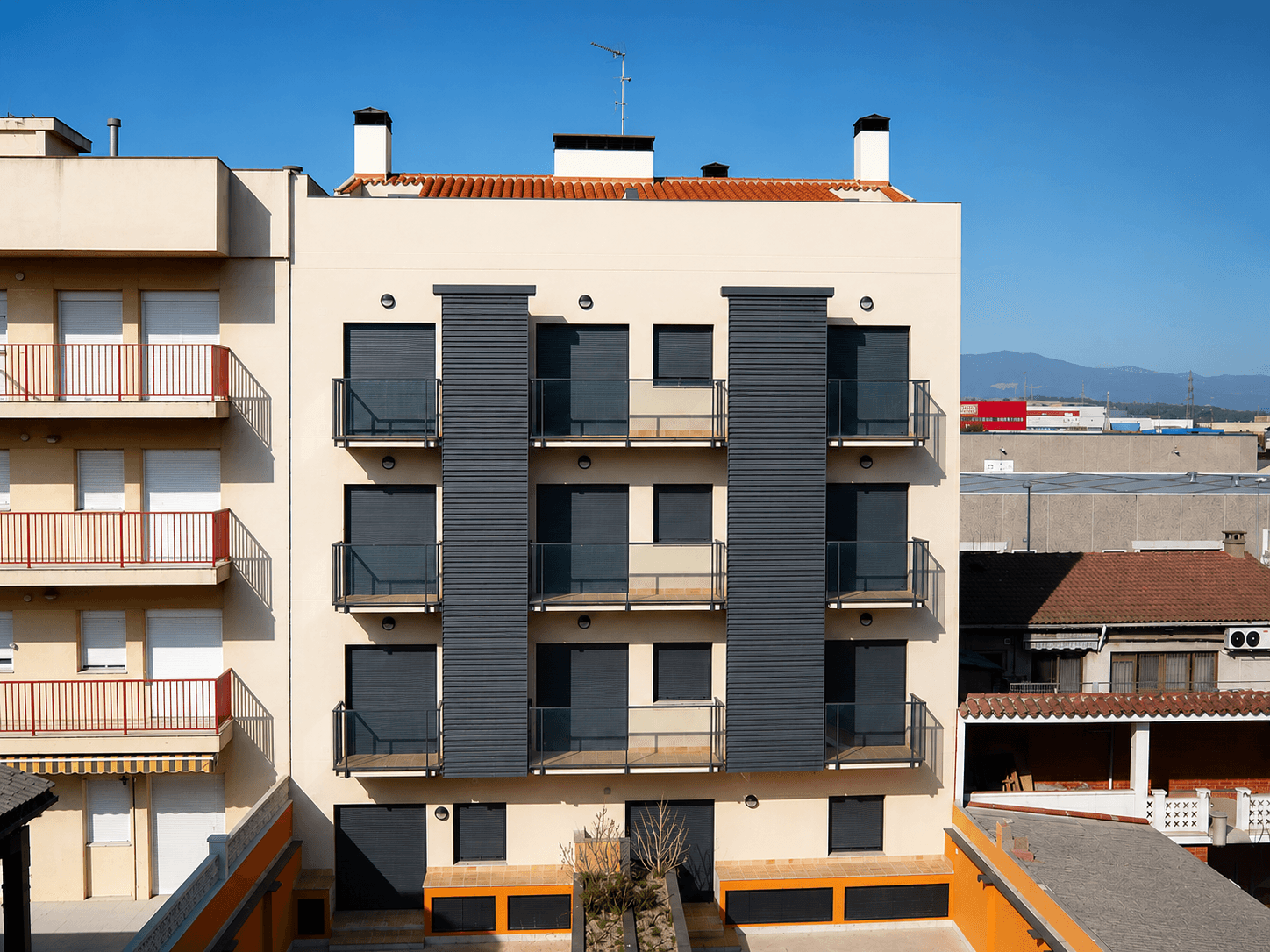 Bloque de cuatro plantas de fachada beige con paños oscuros y balcones de vidrio, con las naves y la montaña al fondo.