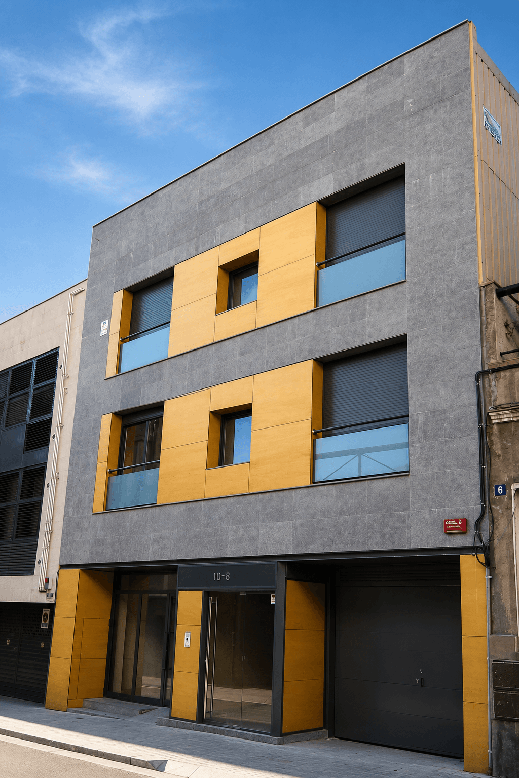 Edificio de tres plantas con fachada gris y paños amarillos, portal acristalado y puerta de garaje, en una calle estrecha.