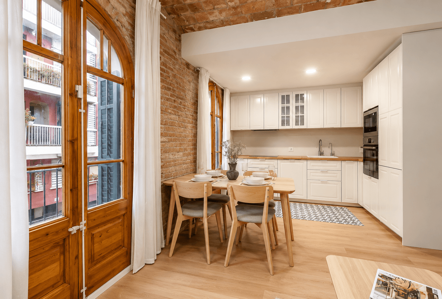 Cocina blanca abierta al comedor, con mesa y sillas de madera clara, pared de ladrillo visto y balconera de arco con carpintería de madera.