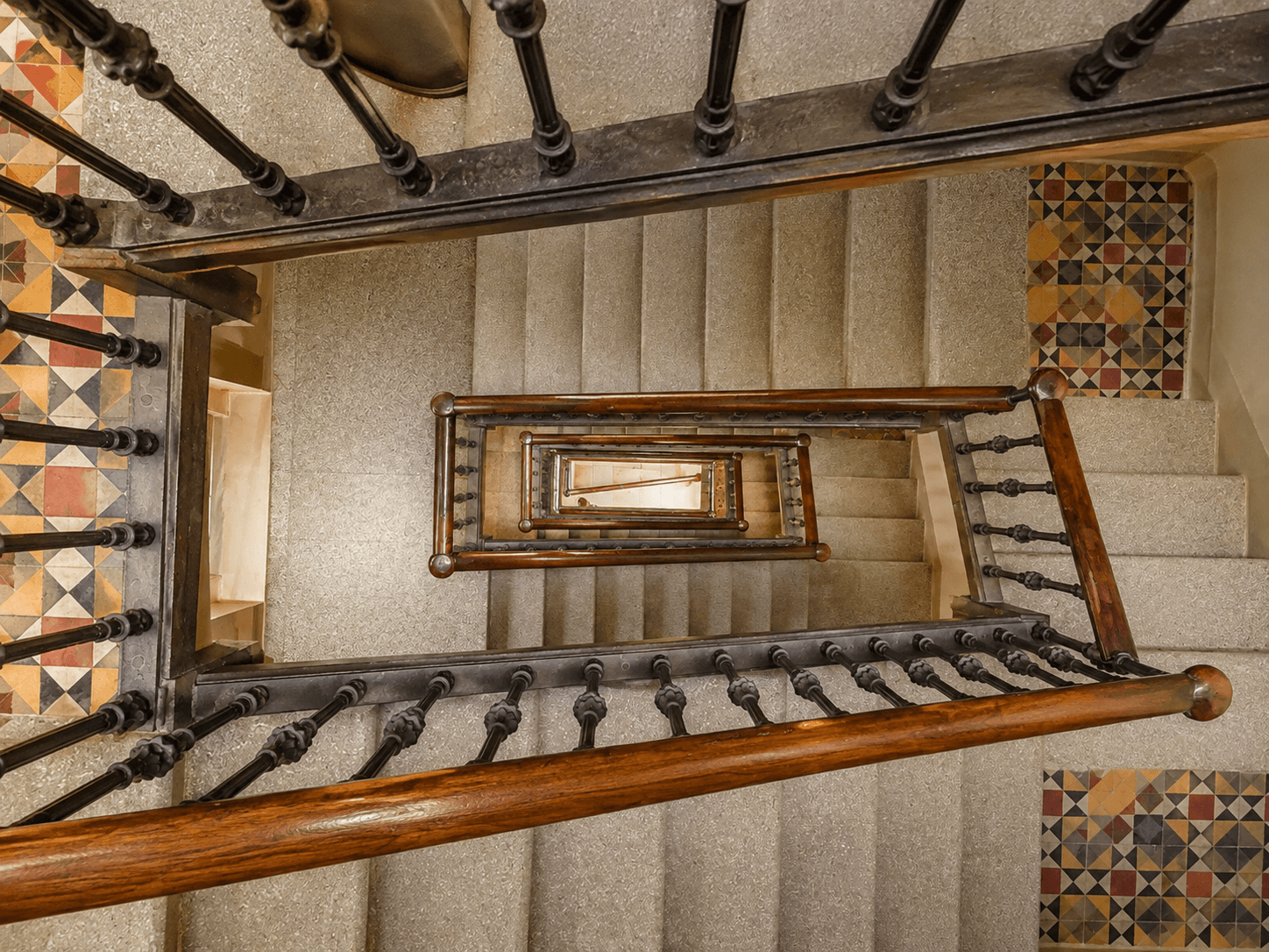 La escalera comunitaria del edificio vista desde arriba, con peldaños de granito, barandilla de hierro con pasamanos de madera y rellanos de mosaico hidráulico original.