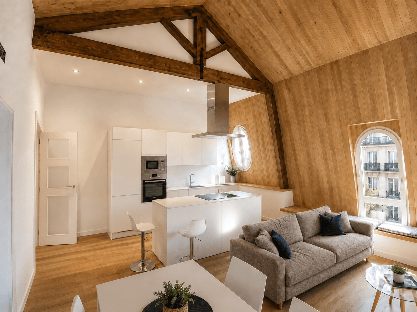 Salón de un ático bajo cubierta a dos aguas, con las cerchas de madera vistas, el faldón forrado de madera, cocina blanca con isla y una ventana de arco sobre la calle.
