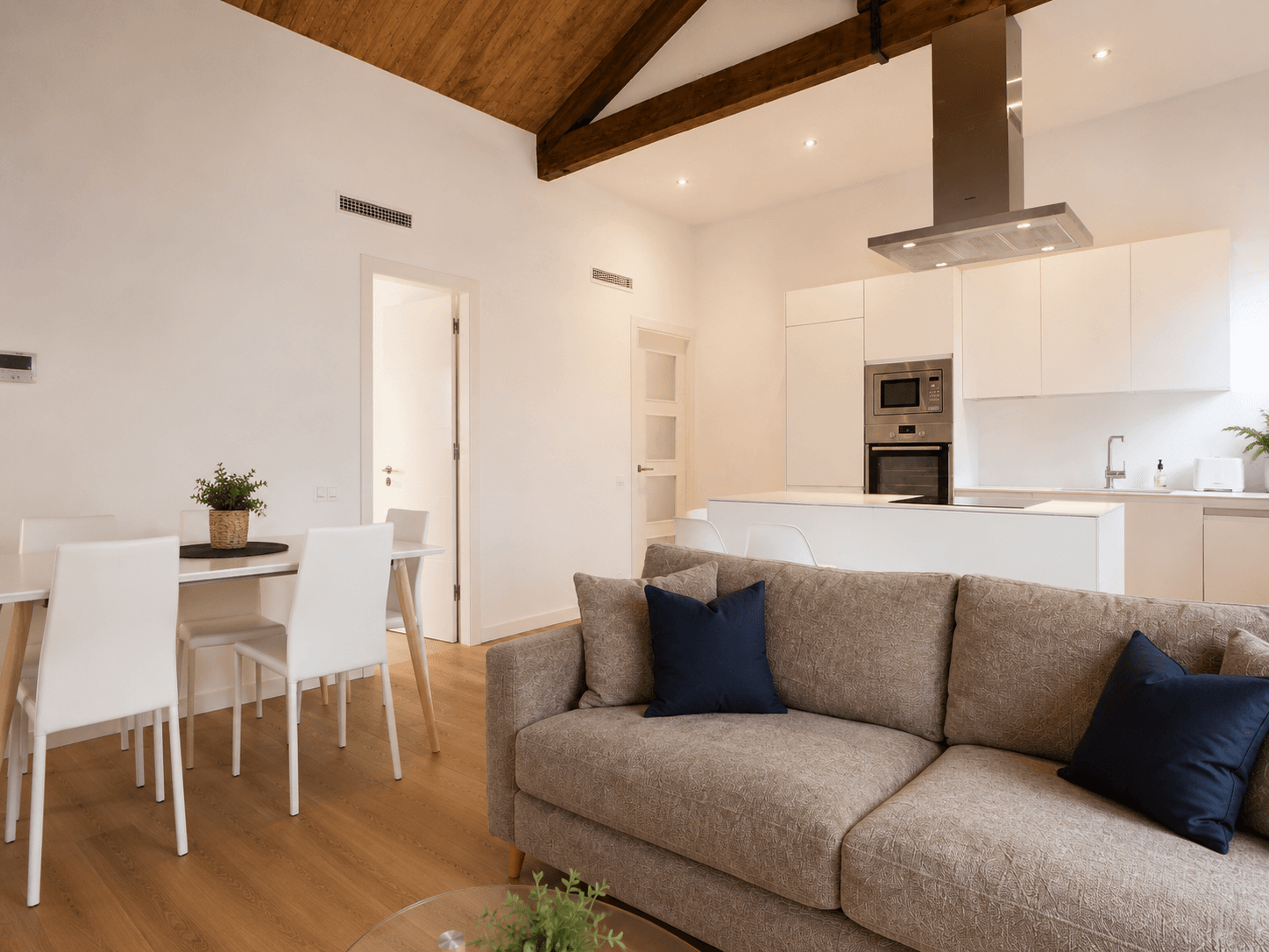 El mismo salón desde el otro extremo: sofá gris, mesa de comedor blanca y la cocina con isla y campana de acero al fondo, bajo la viga de madera.