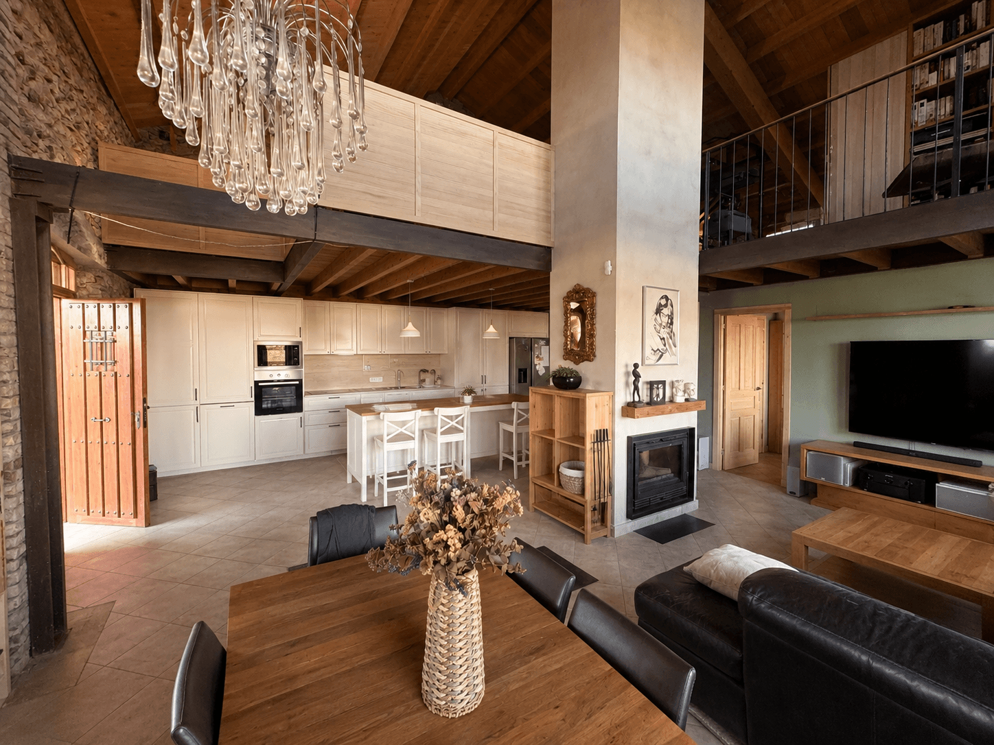 Salón a doble altura con altillo, viguería de madera y cocina blanca con isla al fondo.