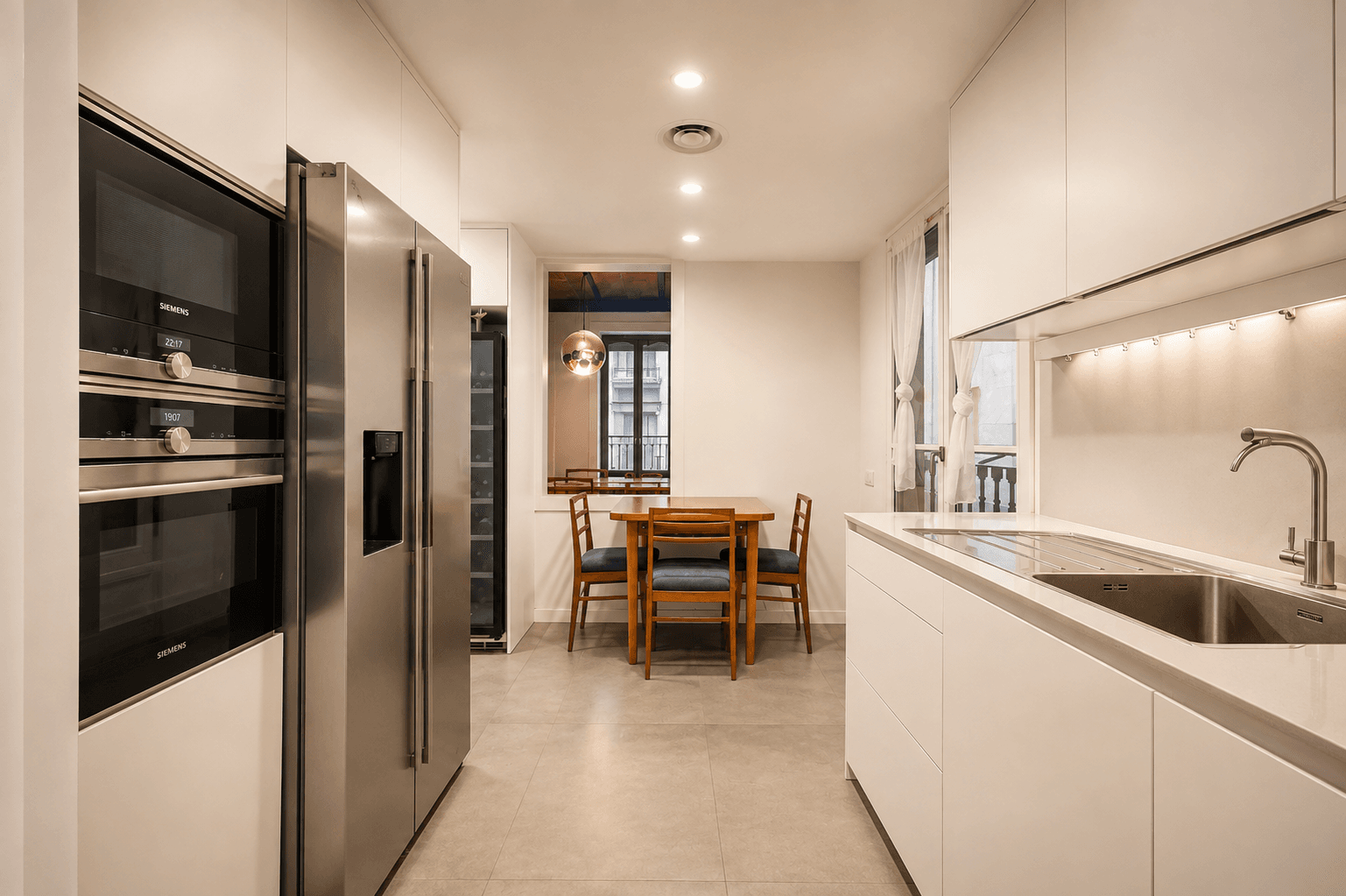 Cocina blanca sin tiradores, con hornos y nevera de acero integrados, encimera continua y un office con mesa de madera junto al balcón.