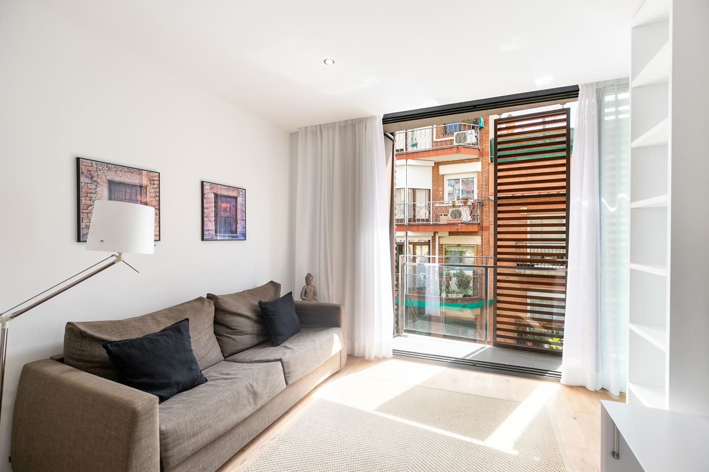 Salón claro con sofá gris, estantería empotrada blanca y ventanal corredero a un balcón con celosía de madera.