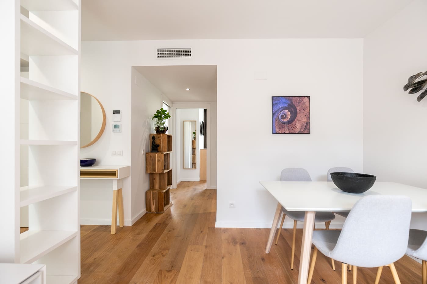 Comedor blanco con mesa y sillas claras, estantería empotrada y pasillo que se ve hasta el fondo de la casa.