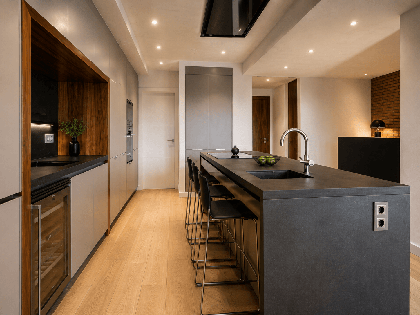 Cocina alargada con isla de encimera de piedra oscura y taburetes, frentes de madera, vinoteca empotrada y suelo de madera clara.