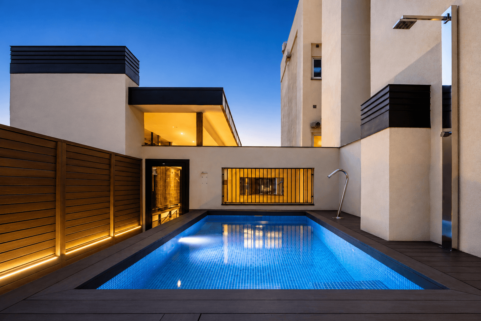 Piscina estrecha entre medianeras al anochecer, con tarima de madera, celosía retroiluminada y la casa encendida al fondo.