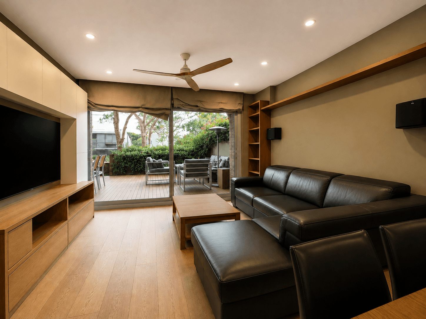 Salón alargado con mueble de televisión a medida, sofá de piel oscuro y suelo de madera clara, abierto a una terraza con porche y jardín.